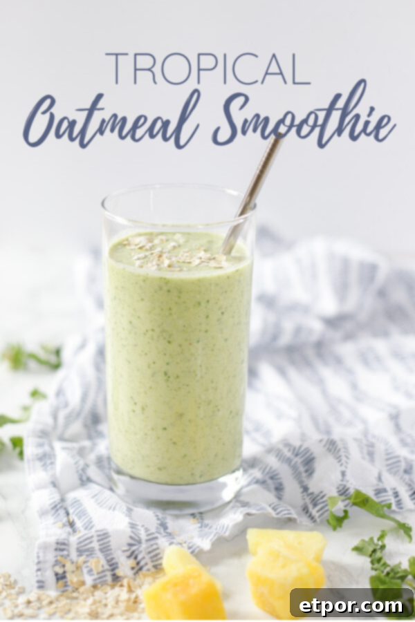 A vibrant green tropical oatmeal smoothie served in a clear glass with a metal straw, garnished with oats on top, surrounded by fresh kale, mango slices, and pineapple chunks on a blue and white patterned napkin.