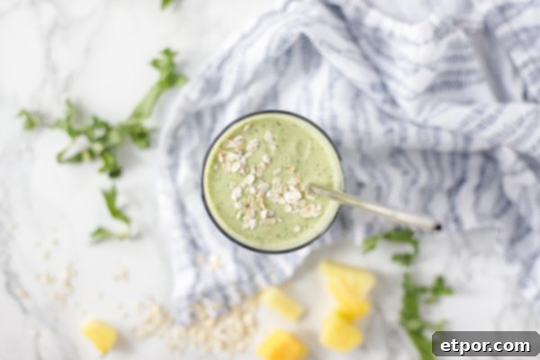 An overhead shot showcasing a tropical oatmeal smoothie in a glass, topped with a sprinkle of oats and a metal straw. The glass rests on a blue and white patterned napkin, with fresh kale, mango slices, and pineapple chunks artfully arranged around it.