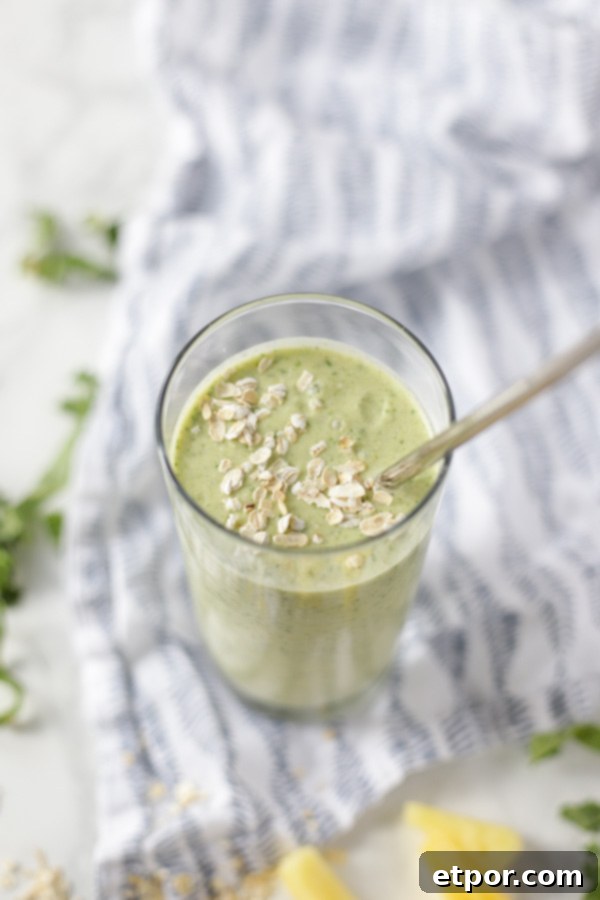 A top-down view of a glass filled with a vibrant green tropical oatmeal smoothie, featuring a metal straw. The glass is positioned on a charming blue and white patterned napkin, artfully surrounded by fresh kale leaves and juicy pineapple chunks.