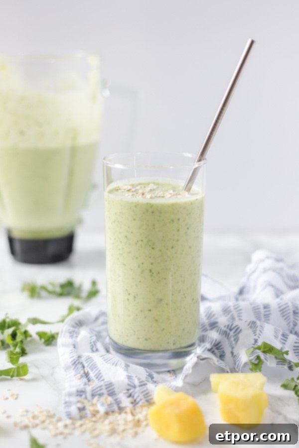 A glass brimming with a vibrant green tropical oatmeal smoothie rests on a blue and white napkin, with the blender jar, still containing some smoothie, visible in the soft-focus background.