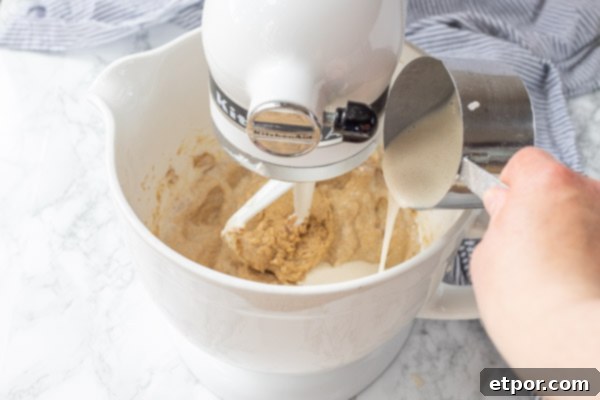 Cultured Chocolate Chip Cookies 6 Sourdough starter, a large egg, and vanilla extract being added to the creamed butter and sugar mixture in a white ceramic mixing bowl on a stand mixer.