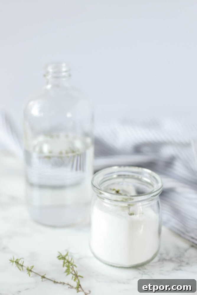 toilet bowl cleaner in a small glass jar and a bottle of vinegar in a tall glass bottle on a white countertop with fresh herbs