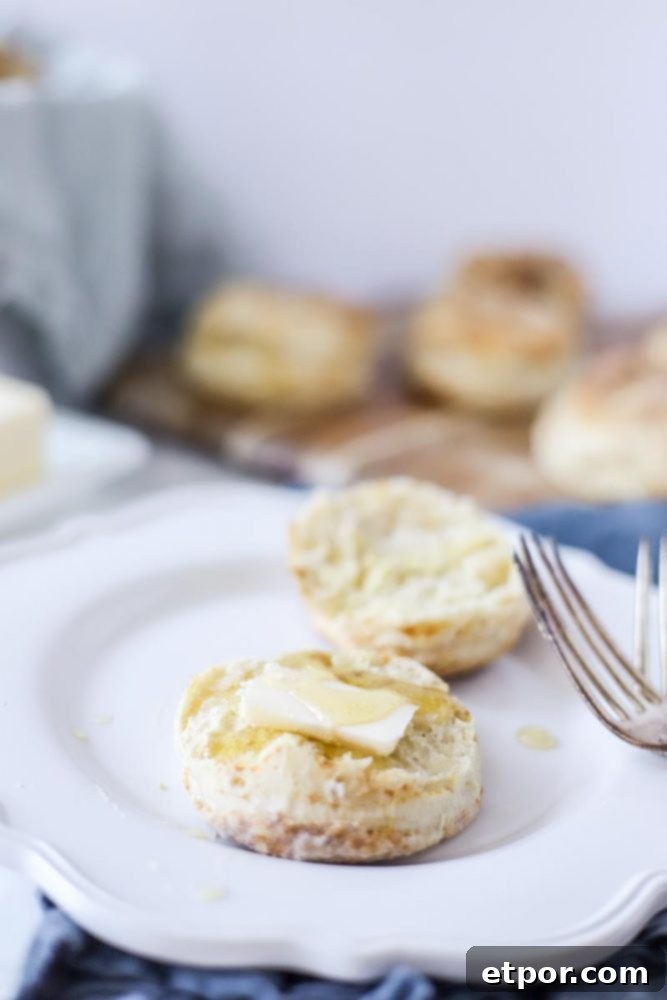 sourdough biscuit cut in half with a pat of butter and a drizzle of honey on a white plate. More biscuits in the background on a cutting board