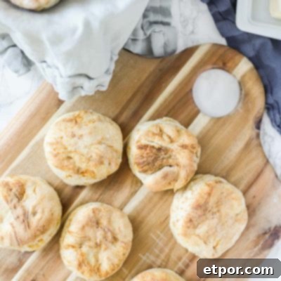 sourdough biscuits on a wood cutting board with a blue napkin lined bowl with more biscuits on the back left corner