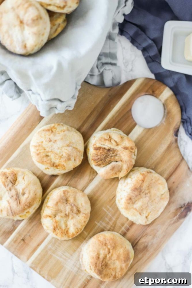 sourdough biscuits on a wood cutting board with a blue napkin lined bowl with more biscuits on the back left corner