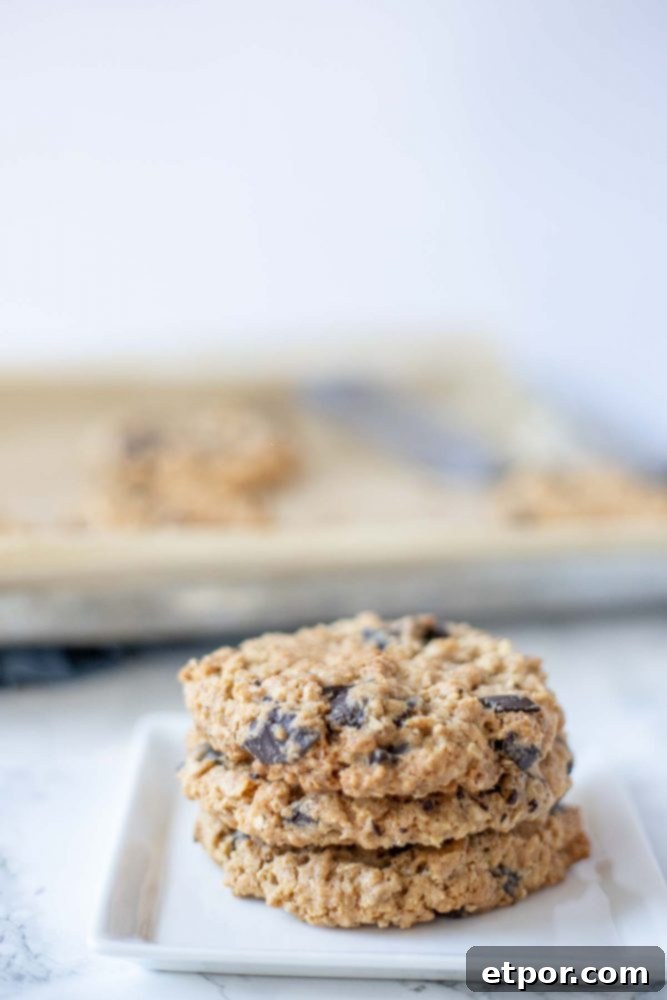 three chocolate chip oatmeal cookies stacked up on a white plate with a pan of cookies in the background