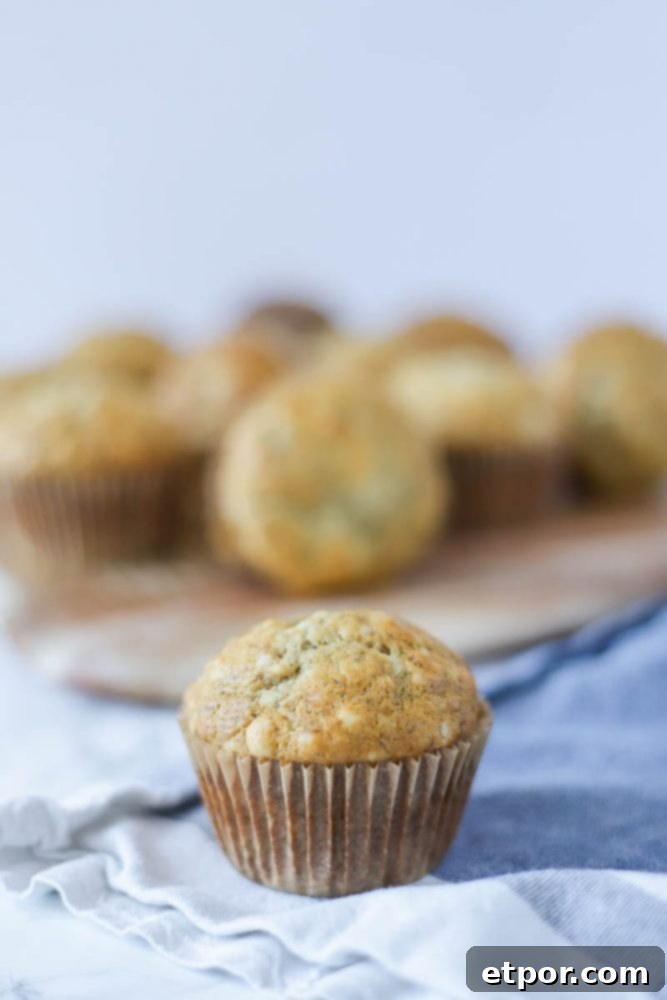 A single sourdough banana muffin rests on a white and blue striped towel, with more muffins blurred in the background on a wooden board.
