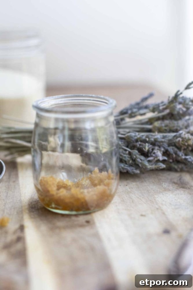 Whip Up Your Own Lip Scrub 4 jar of lip sugar scrub on a wood countertop with a bundle of lavender and a glass of sugar in the background