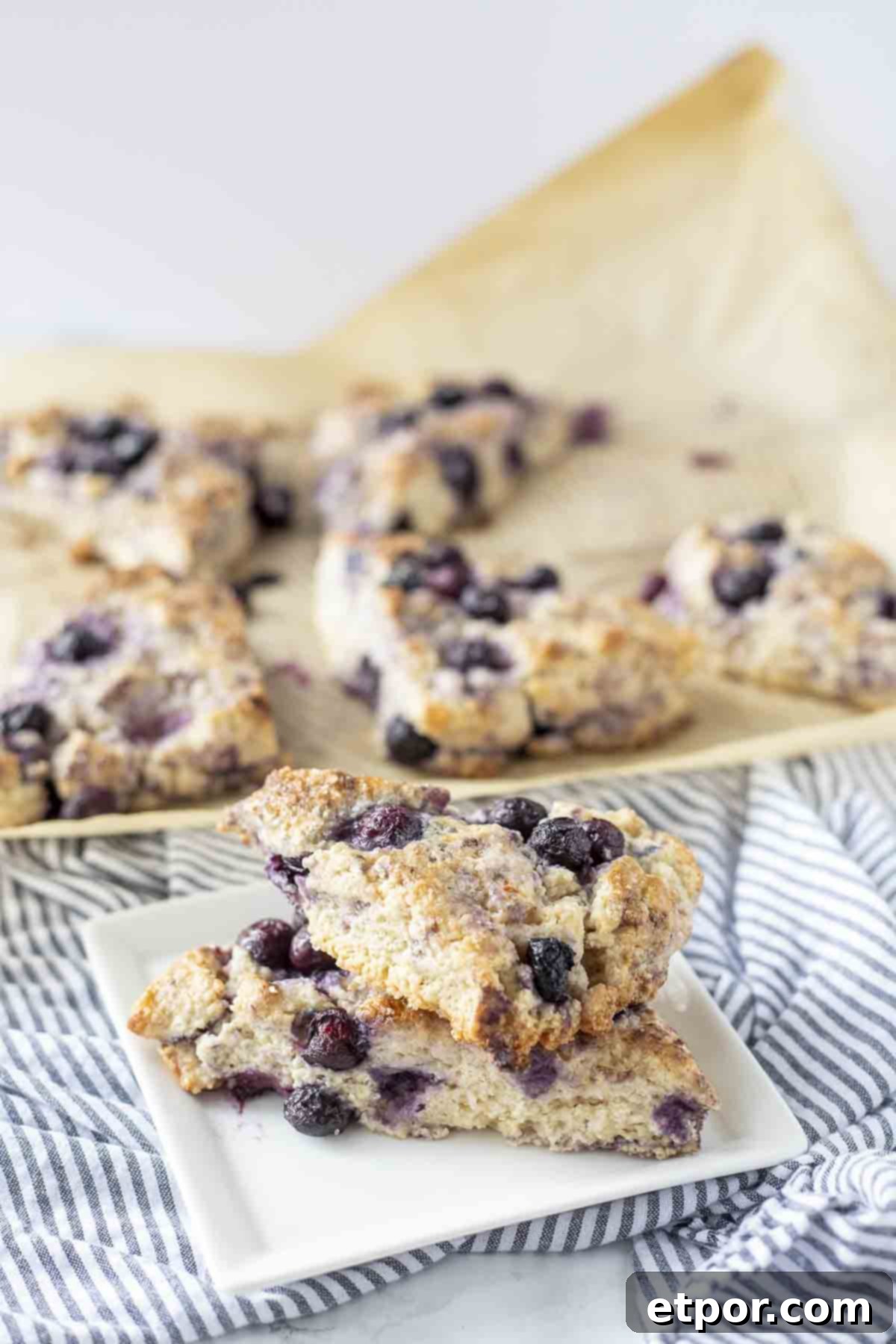 Effortless Sourdough Blueberry Scones 3 Two blueberry sourdough scones stacked on a white plate, placed on a white and blue striped towel, with more freshly baked scones blurred in the background, highlighting the rustic charm.