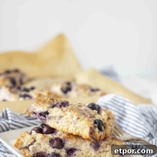 Effortless Sourdough Blueberry Scones 8 Two golden-brown sourdough blueberry scones, lightly glazed, stacked on a white plate, showcasing their fluffy interior and juicy blueberries.