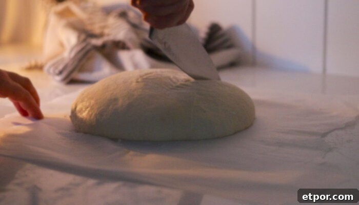 A knife scoring the top of a sourdough boule, prepared for baking.