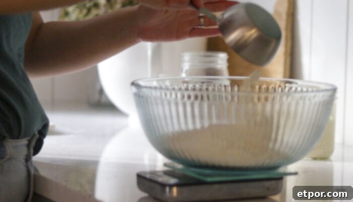 Glass bowl on a kitchen scale. A person is adding flour to the bowl.