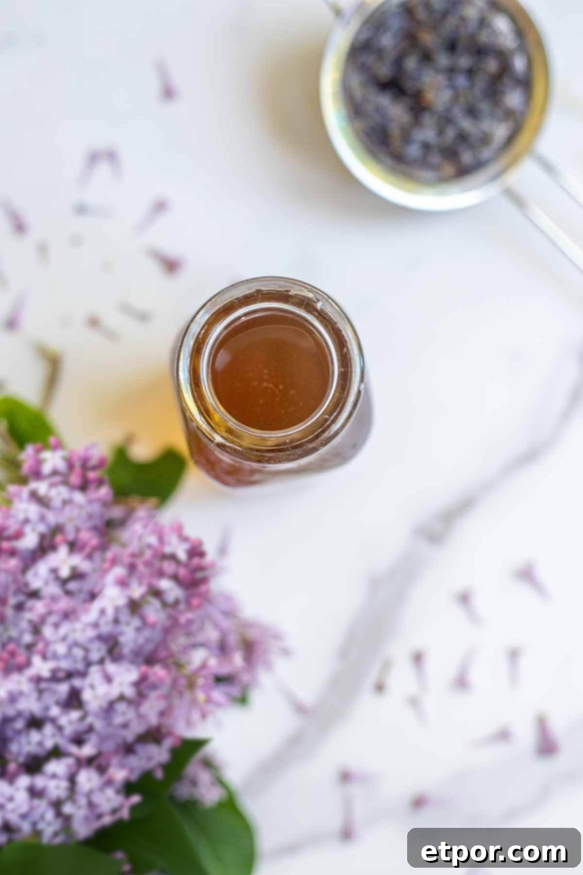 overhead photo of lilac syrup in a jar with fresh lilac flowers and steeped strain flowers surrounding the jar.