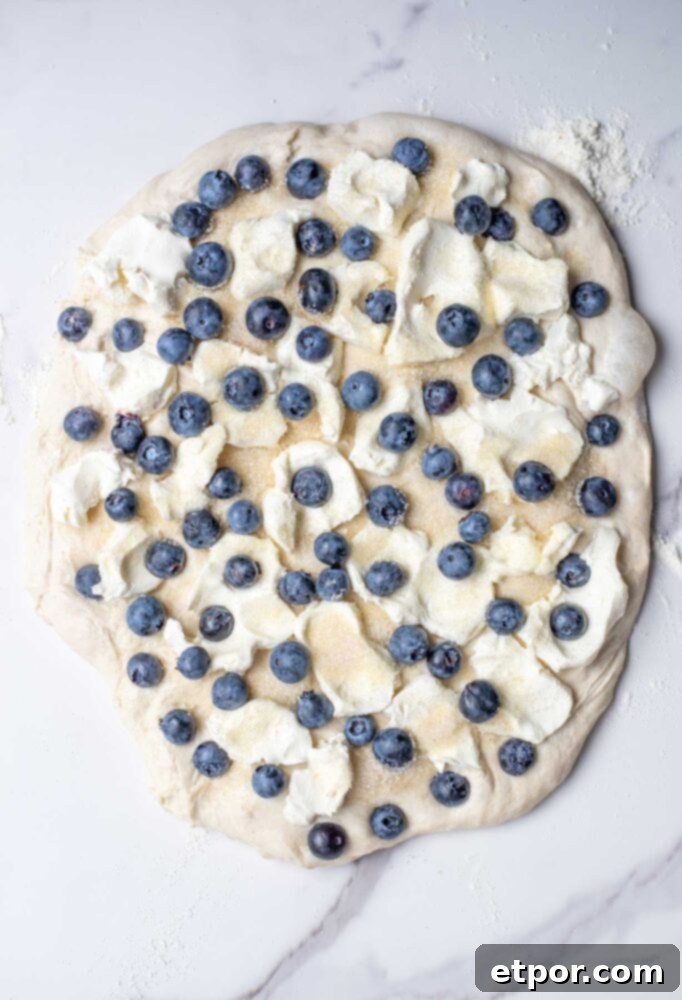 Sourdough bread dough patted out into a large circle and topped with cream cheese, blueberries and sugar.