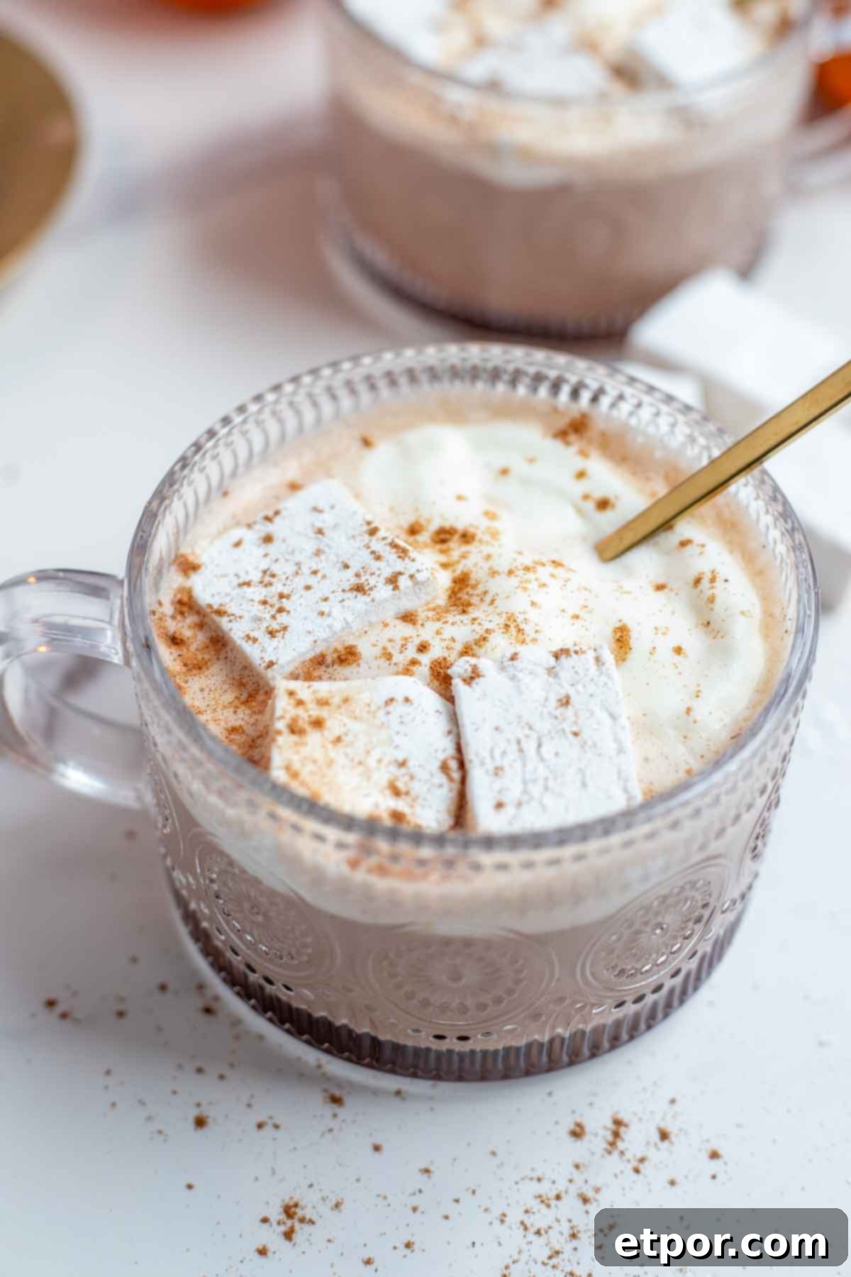Close up picture of pumpkin spice hot chocolate topped with whipped cream, marshmallows, and pumpkin spice with a gold spoon. More hot chocolate in th background.