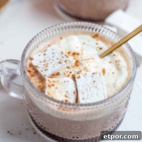 Close up picture of pumpkin spice hot chocolate topped with whipped cream, marshmallows, and pumpkin spice with a gold spoon. More hot chocolate in th background.