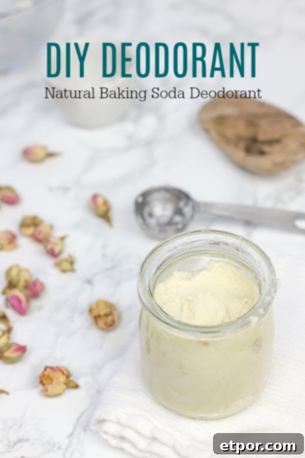 Homemade DIY deodorant in a clear glass jar, placed on a white washcloth with scattered rosebuds and a stainless steel measuring spoon nearby.