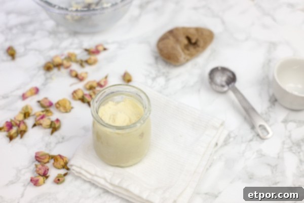 A glass jar of DIY deodorant sits on a white washcloth, surrounded by delicate rosebuds, highlighting its natural and simple ingredients.