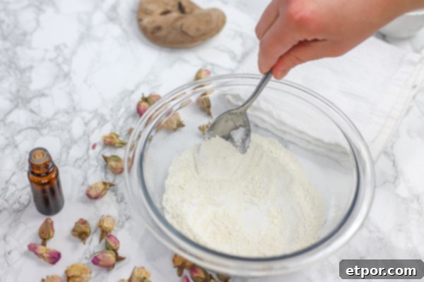 Glass bowl containing dry ingredients for DIY deodorant being stirred, with scattered dried rose petals for aesthetic.