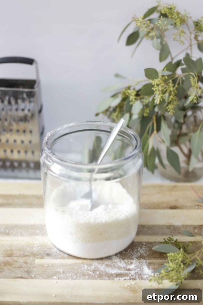 homemade laundry detergent in a jar with a spoon. The jar is on a wood cutting board with a bouquet of eucalyptus and a cheese grater in the background.