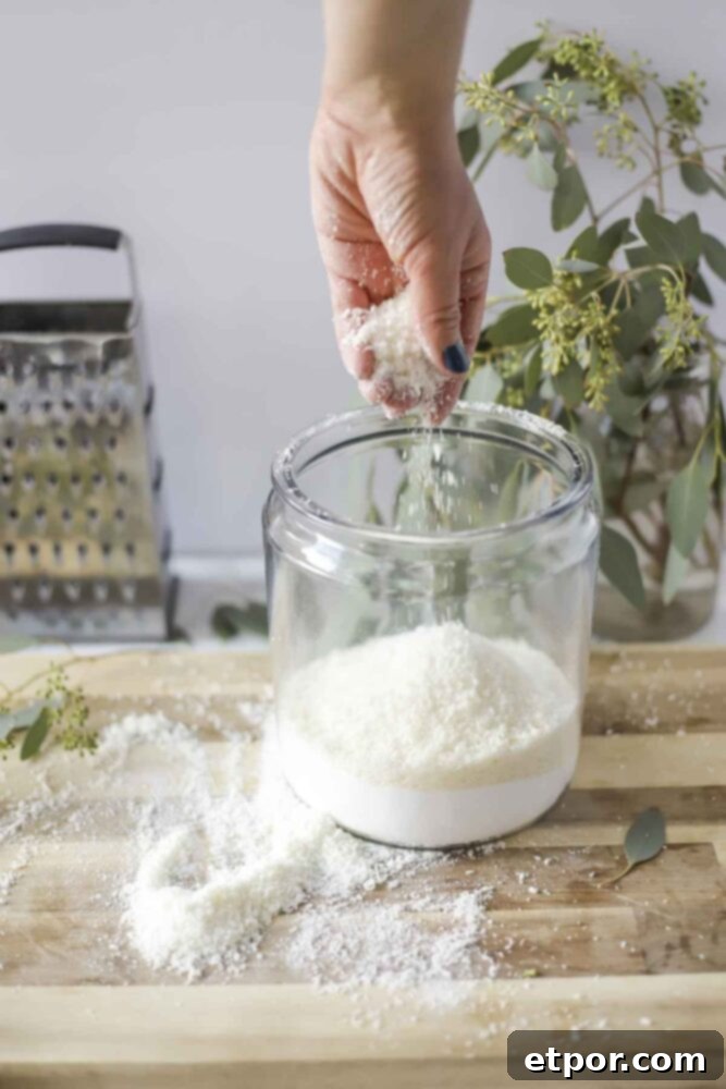 hand adding grated soap to a large class container of borax and washing soda to make homemade laundry detergent