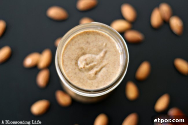 A jar of creamy homemade almond date butter with a spoonful being drizzled back into the jar, surrounded by scattered almonds and dates.