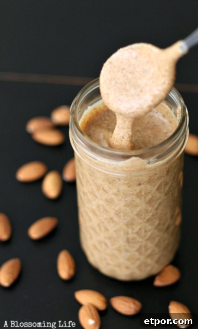 A spoon drizzling homemade almond butter into a mason jar, with roasted almonds and dates scattered on a wooden counter.