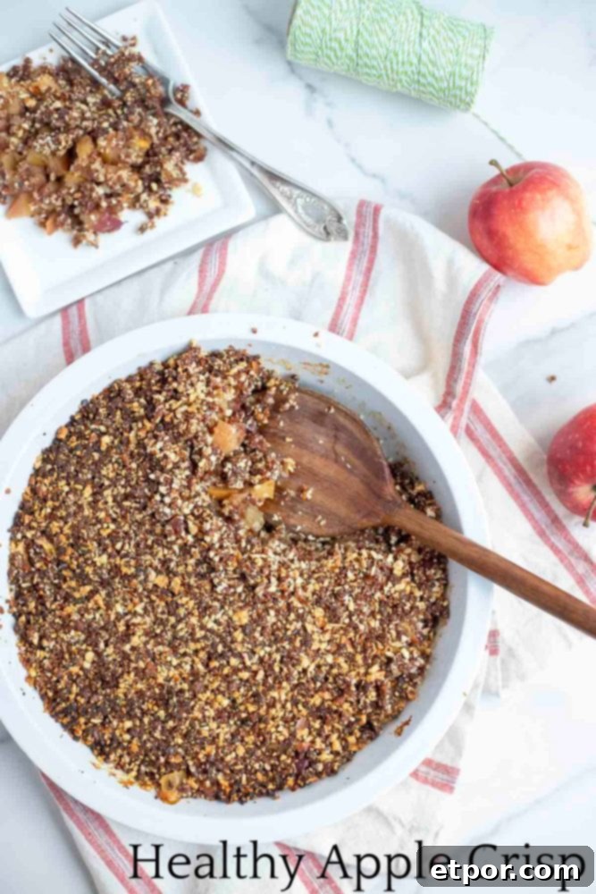 overhead photo of a healthy apple crisp in a pie plate with a wooden spoon in the dish. The pie dish is on a white and red stripped towel with a plate of crisp and apples in the background