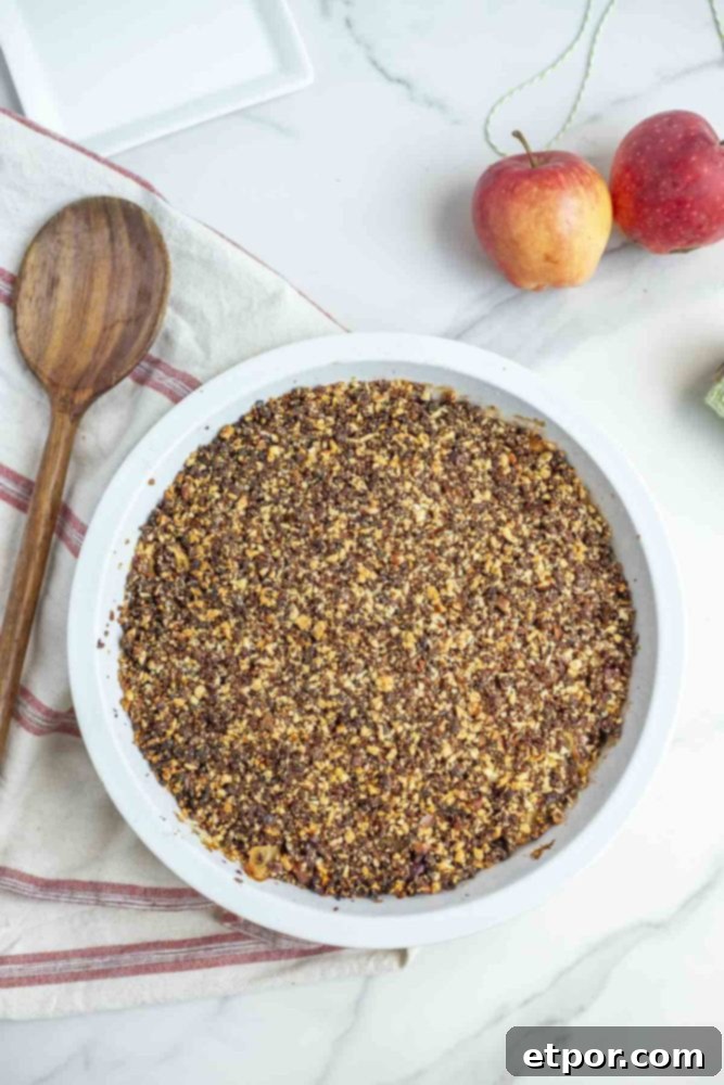 overhead photo of gluten free apple crisp in a pie plate on a white and cream stripped towel with a wooden spoon