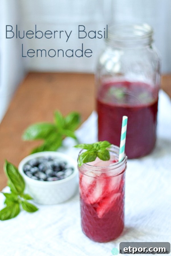 A tall glass of homemade blueberry basil lemonade garnished with a sprig of fresh basil and a teal straw, sitting on a white kitchen towel with scattered blueberries.