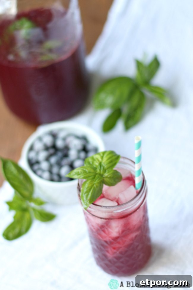 Blueberry Lemonade in a mason jar topped with a sprig of basil. A white dish of frozen blueberries sits behind it, highlighting the fresh ingredients.