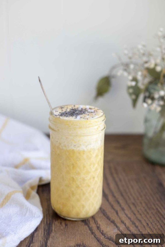 Spiced Pumpkin Pie Bliss Smoothie 7 healthy pumpkin smoothie in a glass jar with a spoon in the smoothie. The jar is on a wood countertop with a white and yellow towel and a jar of flowers int he background