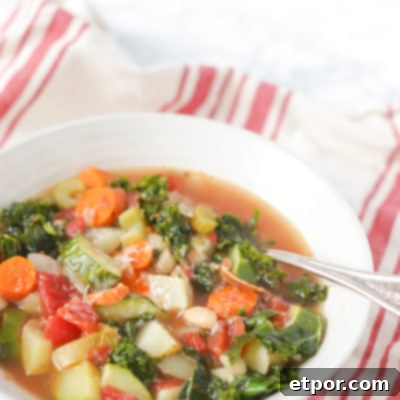 A hearty bowl of minestrone soup with bacon, kale, potatoes, and other vegetables in a rich chicken stock, served with an antique spoon on a red and tan napkin.