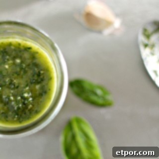 Homemade vegan pesto in a jar, surrounded by fresh basil leaves and garlic cloves.