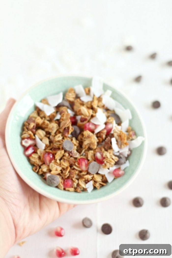 Whip Up Incredible Homemade Granola 5 A hand gently holding a teal bowl brimming with homemade granola, generously topped with an appealing mix of chocolate chips, ruby-red pomegranate seeds, and delicate coconut shavings. More chocolate chips and pomegranate seeds are artfully scattered around the bowl.