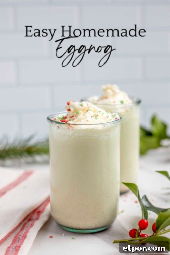 two glasses of homemade eggnog topped with whipped cream and sprinkled with christmas sprinkles on a marble countertop surrounded by greenery and a red and cream towel