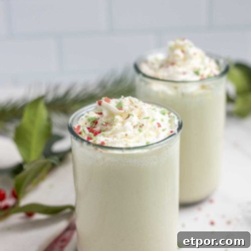 two jars of homemade eggnog topped with whipped cream and red, green, and white sprinkles on a marble countertop with greenery in the background
