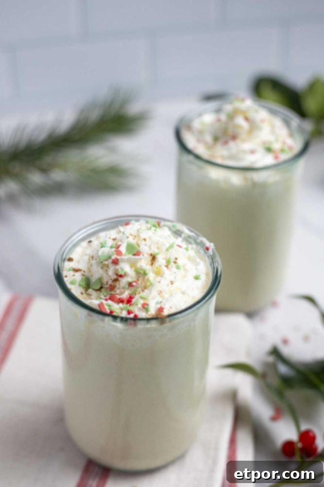 two glasses of homemade eggnog topped with whipped cream and sprinkles. The glasses are on a white and cream strolled towel and surrounded by greenery