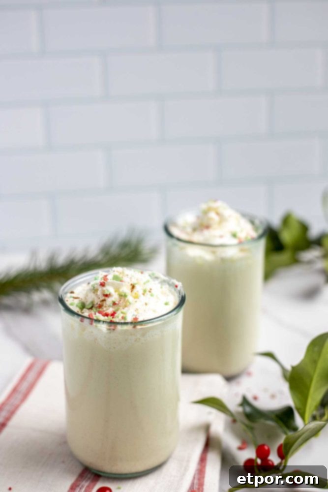 two glasses of easy homemade eggnog on a cream and red stripped towel with holly and greenery surrounding the glasses