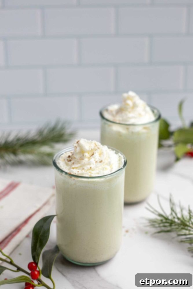 two large glasses of homemade eggnog with whipped cream on marble countertop with a red and white stripped towel to the right and greenery surrounding the glasses