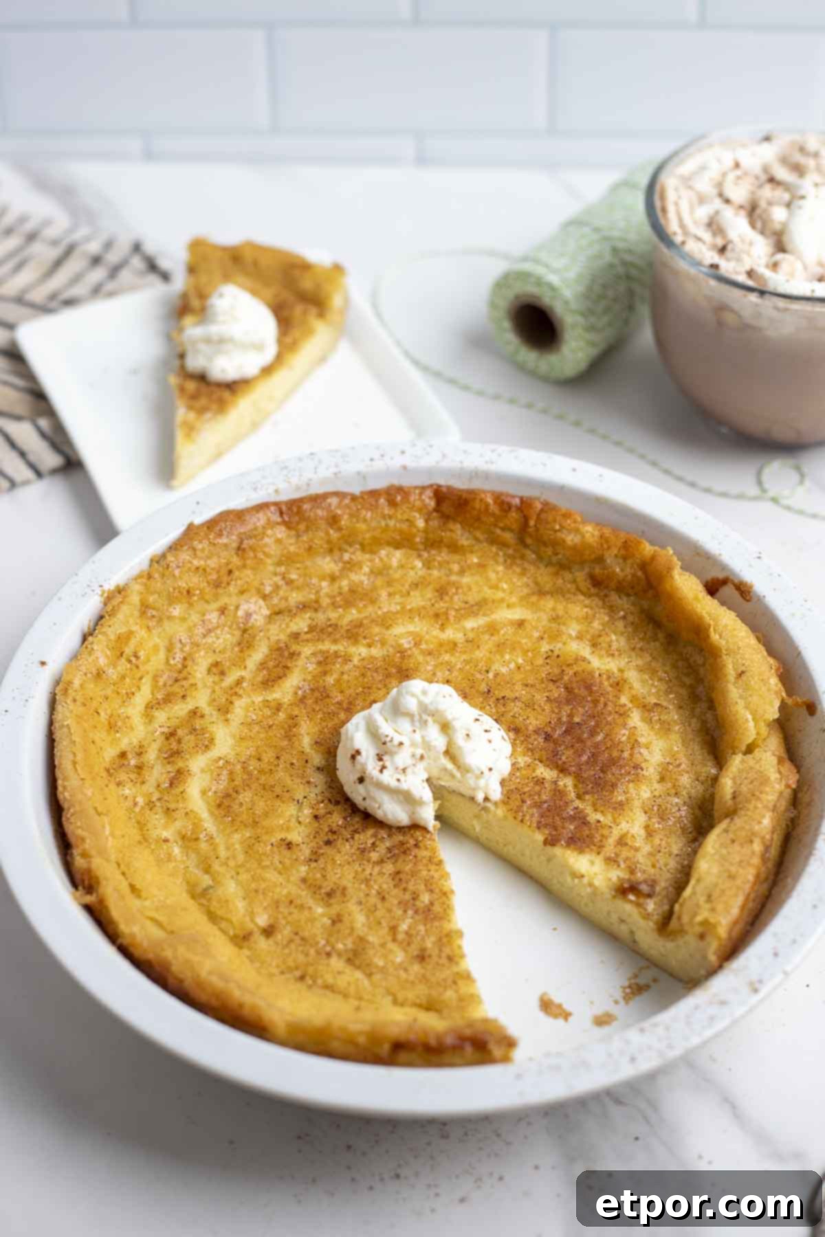 custard pie with a slice taken out and a dollop of whipped cream in a white pie plate. The pie is on a marble counter with a slice of pie, hot chocolate, and green string in the background.