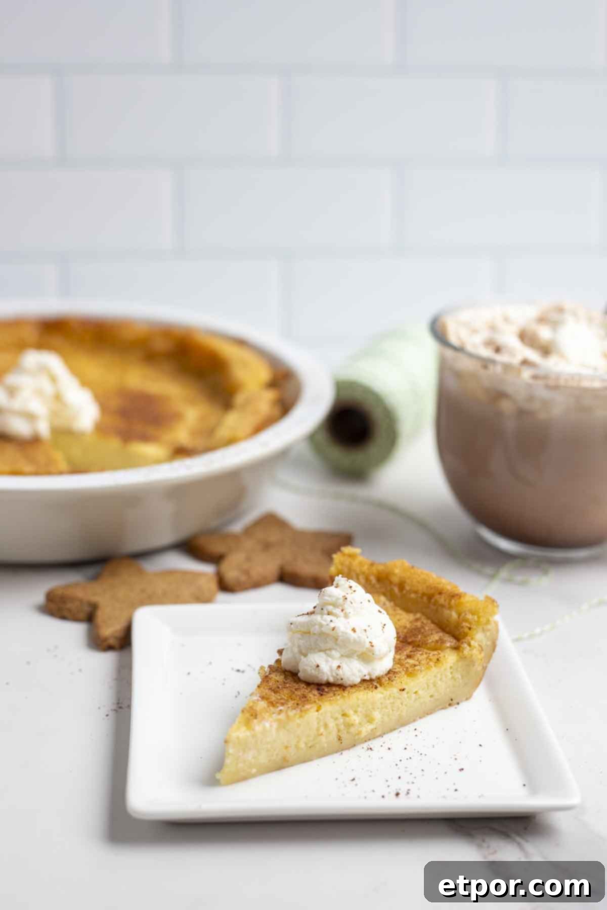 slice of custard pie with a dollop of whipped cream on a white square plate on a marble countertop. The pie plate with pie, hot chocolate, and cookies are in the background.