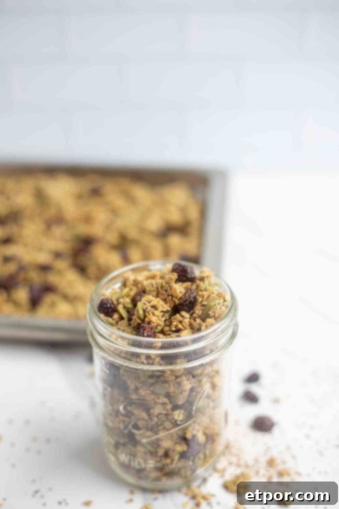 Irresistible Pumpkin Spice Granola 13 jar of pumpkin granola on a marble countertop with oats, and seeds around the jar. A pan of granola is in the background