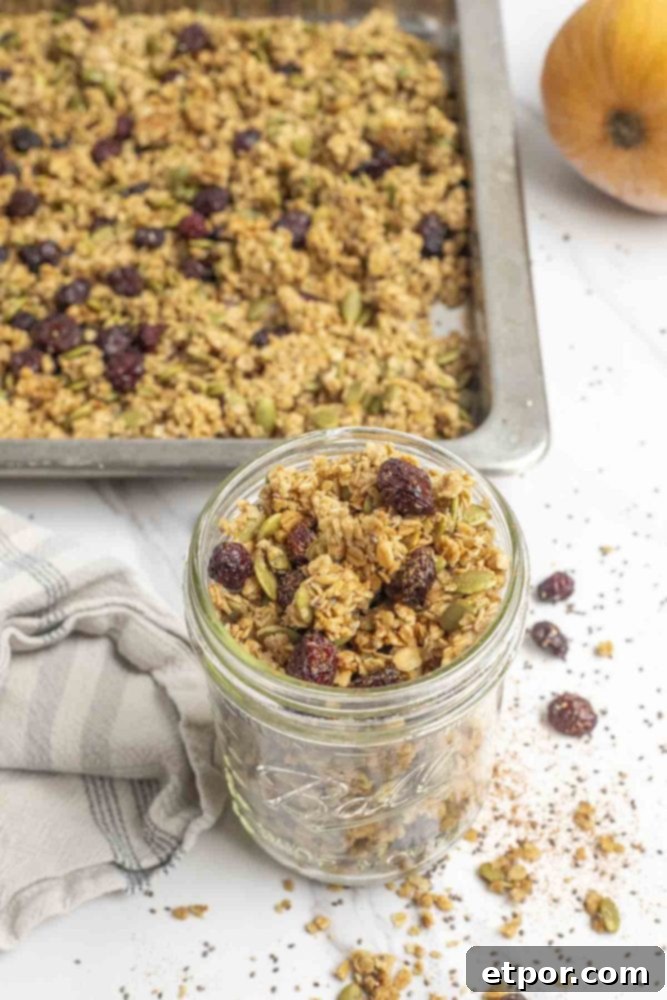 Irresistible Pumpkin Spice Granola 6 Pumpkin spice granola with pumpkin seeds and craisins in a mason jar with ingredients spread around the jar. A pan of granola and a pumpkin are in the background