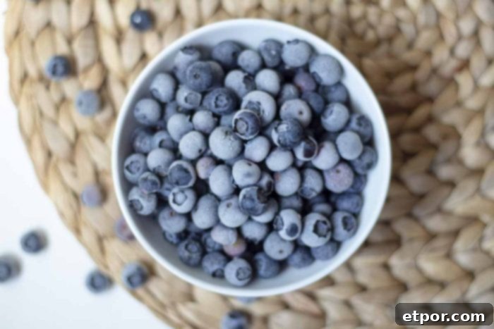 A bowl of vibrant frozen blueberries rests on a woven placemat, surrounded by fresh blueberries, showcasing the bounty of the harvest.