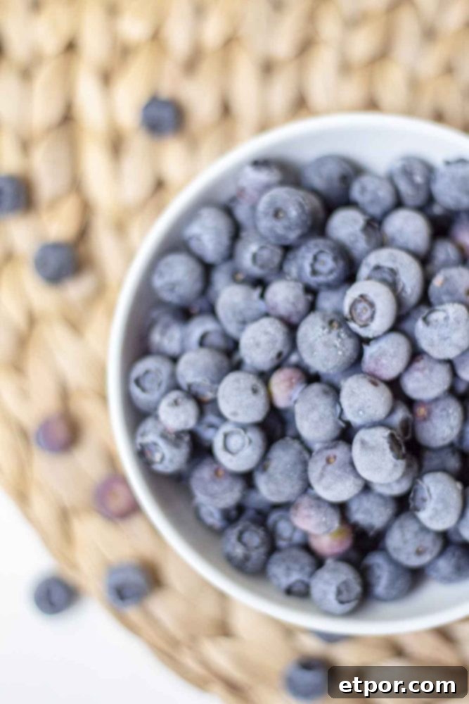 Overhead photo of vibrant frozen blueberries in a white bowl, resting on a woven mat, surrounded by a scattering of fresh berries.