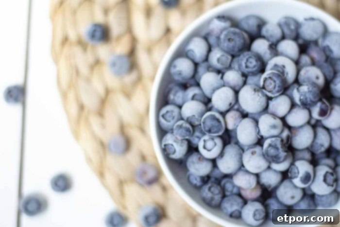Overhead photo of frozen blueberries in a white bowl, with more fresh blueberries scattered on a woven mat.