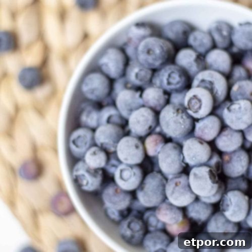 Overhead photo of frozen blueberries in a white bowl, resting on a woven mat, surrounded by a scattering of fresh berries.