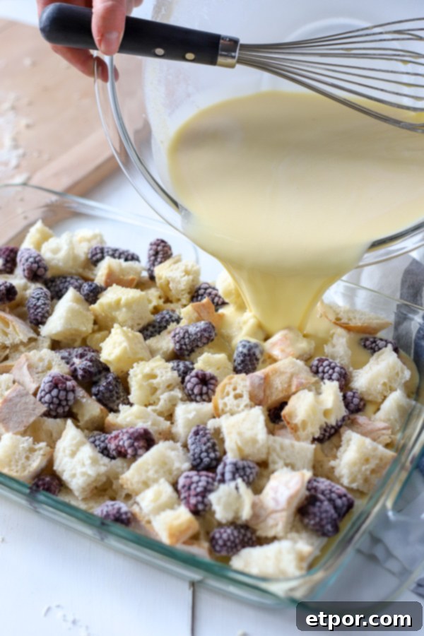 Rich custard being poured over layers of bread and fresh blackberries in a glass baking dish