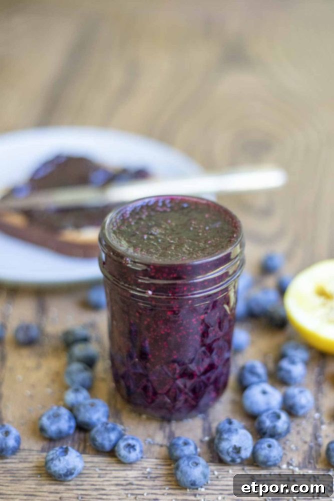 jar of blueberry chia jam in a small mason jar surrounded by fresh blueberries. A plate with toast with jam and a knife in the background